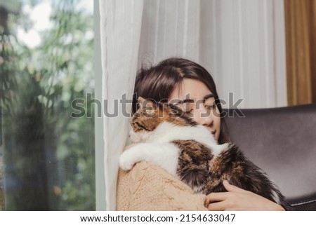Asian woman holding cat over her shoulder with clossed eyes and smile of happiness beside glass window and white curtain while cat looking out of window. Royalty-Free Stock Photo #2154633047