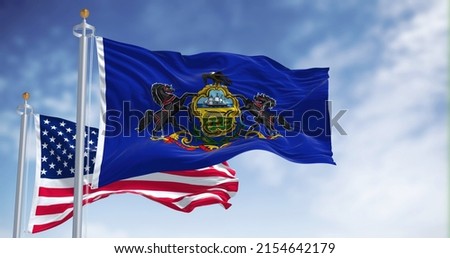 The Pennsylvania state flag waving along with the national flag of the United States of America. In the background there is a clear sky. Pennsylvania is a U.S. state in the northeast Royalty-Free Stock Photo #2154642179