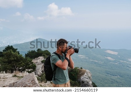 A young male tourist with a backpack climbed to the top of the mountain and takes pictures on the peak from a height. The concept of active life and hiking.
