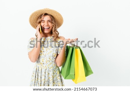 Joyful young woman winking while holding shopping bags and talking on mobile phone, white studio background