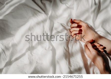 Elegant hands lie on the white bed sheet in the sunlight. Bed with white linens. The concept of a good morning, stress relief, self-care, relaxation and time for yourself Royalty-Free Stock Photo #2154671255