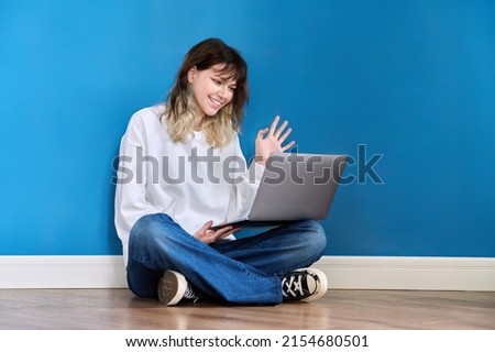 Beautiful teenage female sitting on the floor with laptop on blue background