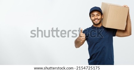 Delivery Concept - Portrait of Happy African American delivery man holding a box package and showing thumps up. Isolated on Grey studio Background. Copy Space. Royalty-Free Stock Photo #2154725175