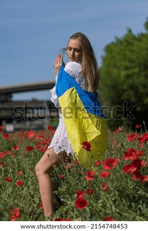 A young blonde Ukrainian woman stands in a field of Red Poppy flowers holding the flag of Ukraine showing her support for the war in her native country of Ukraine