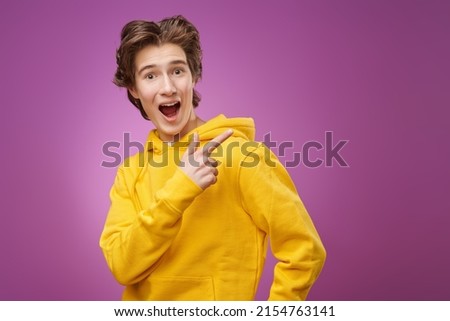 Portrait of a surprised, excited  teenage boy in bright yellow hoodie pointing his index finger away. Purple background with copy space. Adolescents and lifestyle. Youth fashion.  Royalty-Free Stock Photo #2154763141