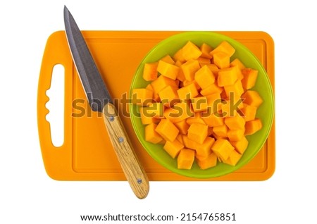 Cubes of raw pumpkin in green glass bowl, kitchen knife on plastic cutting board isolated on white background. Top view