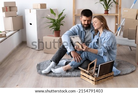 happy young couple sit on floor in casual clothes hugging looking at framed photo. A pile of cardboard boxes and houseplants near a white wall indoors. Moving day. copy space.