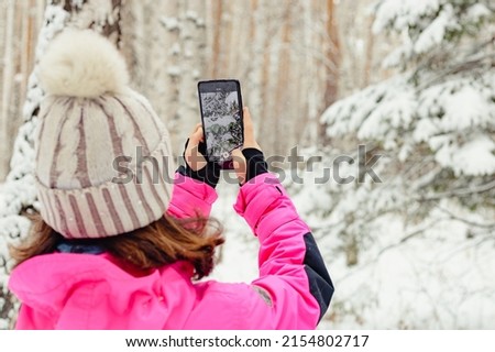 The girl takes pictures of nature on a smartphone. A teenager in bright warm overalls takes a photo of a winter forest on a cell phone camera