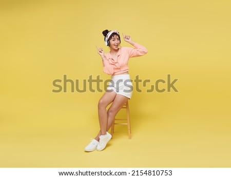 Happy young Asian teen woman smiling listening music in headphones and sitting on chair isolate on bright yellow background.