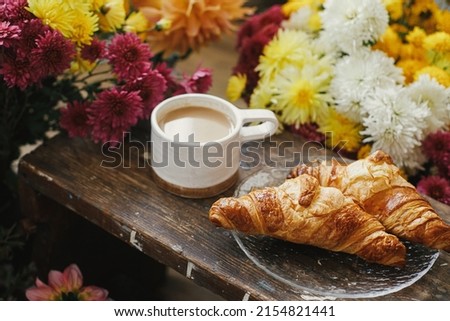 Warm cup of coffee and freshly baked croissants on rustic wooden background with beautiful autumn flowers. Good morning. Stylish mug with cappuccino and croissants on plate, moody image