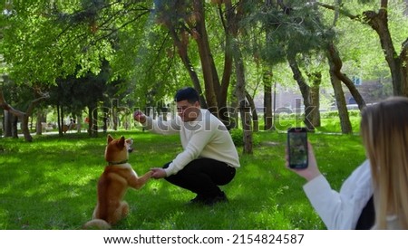 A young couple is sitting in the park on a picnic with a dog. A girl takes pictures of her boyfriend playing with a dog on her phone. Picnic in nature