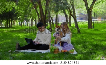 A young couple is sitting in the park on a picnic and stroking their pet dogs on the head. The guy looks at his laptop, and the girl takes a selfie on the phone with the dog. Picnic in nature
