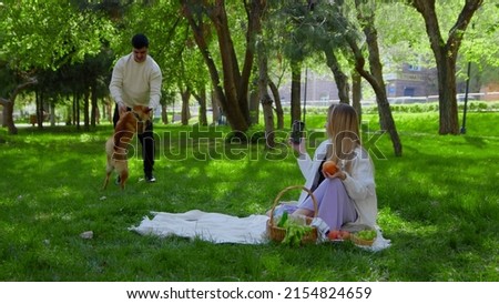 A young couple is sitting in the park on a picnic with a dog. A girl takes pictures of her boyfriend playing with a dog on her phone. Picnic in nature