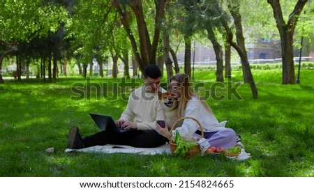 A young couple is sitting in the park on a picnic and stroking their pet dogs on the head. The guy looks at his laptop, and the girl takes a selfie on the phone with the dog. Picnic in nature