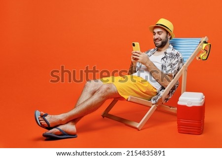Young fun happy tourist man in beach shirt hat lie on deckchair near fridge hold use mobile cell phone isolated on plain orange background studio portrait. Summer vacation sea rest sun tan concept. Royalty-Free Stock Photo #2154835891