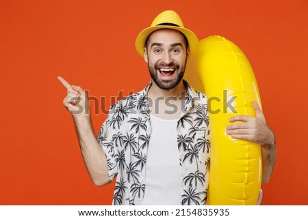 Young surprised tourist man in beach shirt hat hold inflatable ring point index finger aside on workspace isolated on plain orange background studio portrait. Summer vacation sea rest sun tan concept Royalty-Free Stock Photo #2154835935