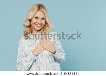 Elderly smiling kind-hearted cheerful caucasian woman 50s wear striped shirt put folded hands on heart isolated on plain pastel light blue color background studio portrait. People lifestyle concept