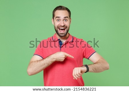 Excited young bearded man guy in casual red pink t-shirt posing isolated on green wall background in studio. People lifestyle concept. Mock up copy space. Pointing index finger on smart watch on hand