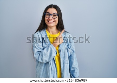 Young hispanic woman standing over blue background cheerful with a smile on face pointing with hand and finger up to the side with happy and natural expression 