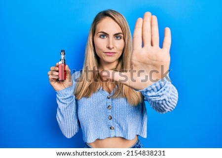 Young caucasian woman holding electronic cigarette with open hand doing stop sign with serious and confident expression, defense gesture 