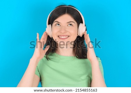 Joyful Young caucasian woman wearing green T-shirt over blue background sings song keeps hand near mouth as if microphone listens favorite playlist via headphones