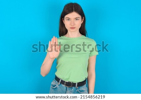 Young caucasian woman wearing green T-shirt over blue background shows stop sign prohibition symbol keeps palm forward to camera with strict expression