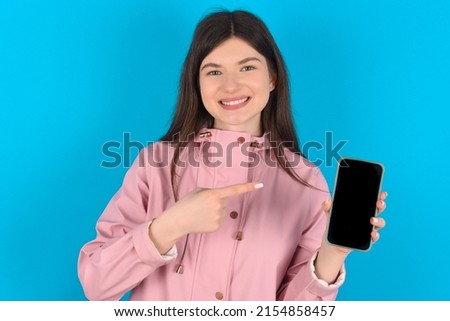Smiling Young caucasian woman wearing pink raincoat over blue background Mock up copy space. Pointing index finger on mobile phone with blank empty screen