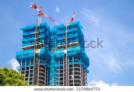 Two multistory buildings, the facade is covered with protective construction mesh on a background of a blue sky. Construction of residential buildings, skyscrapers in big city. Construction cranes. Royalty-Free Stock Photo #2154864753