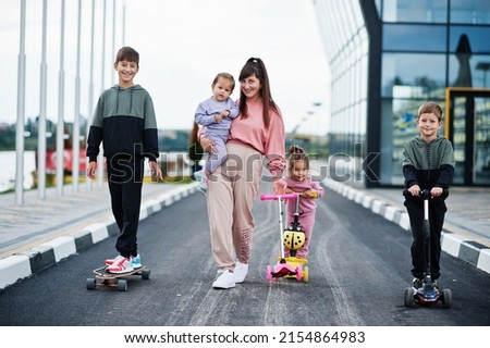 Young stylish mother with four kids outdoor. Sports family spend free time outdoors with scooters and skates.