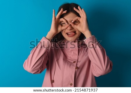 Young caucasian woman wearing pink raincoat over blue background doing ok gesture like binoculars sticking tongue out, eyes looking through fingers. Crazy expression.