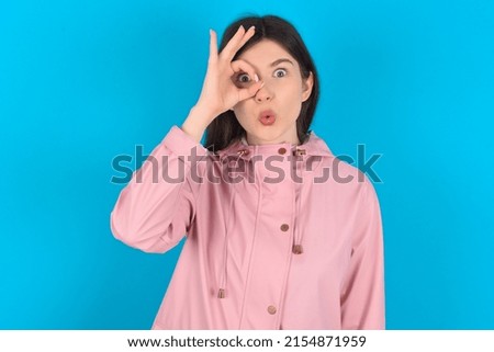 Young caucasian woman wearing pink raincoat over blue background doing ok gesture shocked with surprised face, eye looking through fingers. Unbelieving expression.