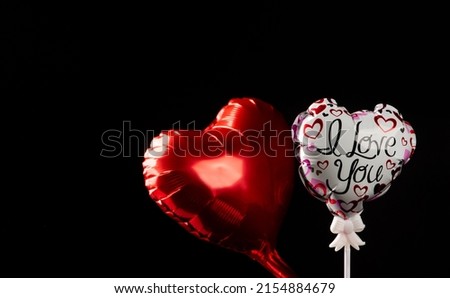 Red heart-shaped balloon and white heart-shaped balloon with the phrase I love you stamped and drawings of hearts, on black background, selective focus on the white balloon.