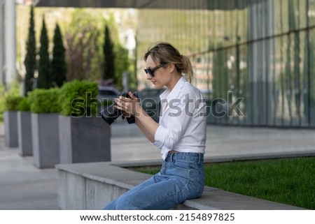 Focused young photographer woman in white shirt blue jeans sit on bench in spring park under the tree outdoors resting using photo camera. Urban lifestyle concept
