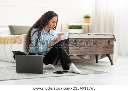 Business And Education Concept. Focused young woman sitting on floor leaning on bed working on laptop writing letter in paper notebook, free copy space. Millennial female studying at home, using pc