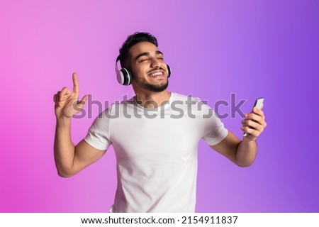 Handsome young Arab man listening to music in wireless headphones, using new smartphone app, closing eyes and dancing in neon light. Attractive middle Eastern guy enjoying favorite playlist or radio Royalty-Free Stock Photo #2154911837
