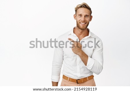 Image of smiling blond male model pointing finger left, showing logo promo aside, wearing white shirt and pants, studio background Royalty-Free Stock Photo #2154928119
