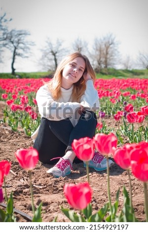 A charming young girl with blond long hair and blue eyes in a light sweater and dark jeans sits against the backdrop of blooming red tulips.