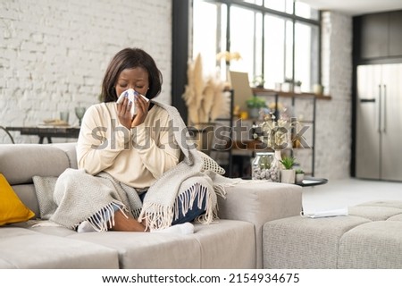 Sick African-American woman sitting covered with blanket on the couch and blowing nose in paper tissue, suffering from seasonal allergy of viral disease, female with runny nose using handkerchief Royalty-Free Stock Photo #2154934675