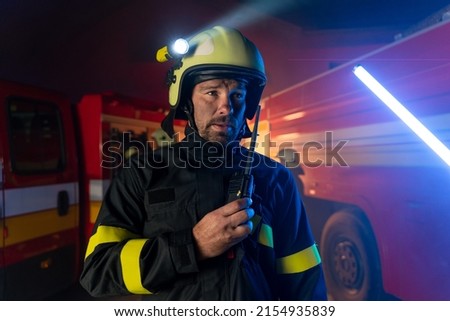 Firefighter talking to walkie talkie with fire truck in background at night.