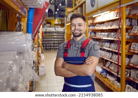 Confident friendly man working in warehouse in hypermarket of building materials. Smiling young male worker in coveralls standing with folded arms in one of aisles between shelves with goods.