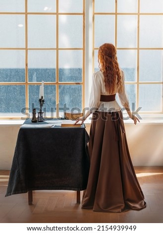 Red-haired woman in vintage dress stands looks at classic window waiting love. Clothing costume countess old style white blouse, brown long skirt. Curly red hair. Redhead girl princess back rear view. Royalty-Free Stock Photo #2154939949