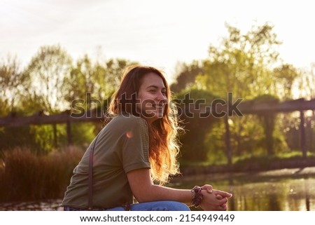 Portrait of a young brunette smiling gir in a summer sunny day with the lake and trees in background. Young girl sitting close the lake with sunlight on her. 