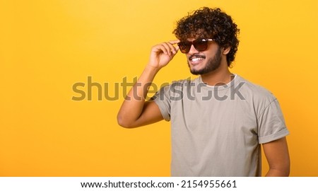 Stylish handsome Indian young curly man wearing casual t-shirt and sunglasses has good mood, enjoying sunny day, posing isolated on yellow background Royalty-Free Stock Photo #2154955661