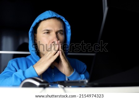 Man in and blue hoodie sitting at laptop with a thoughtful face expression. Concept of solving a difficult task, cybercrime or hacking