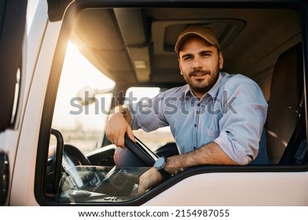 Young truck driver sitting behind steering wheel in a cabin and looking at camera. Royalty-Free Stock Photo #2154987055