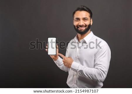 Mock-up concept. Handsome confident bearded Indian man in formal wear showing smartphone with empty screen, points finger at blank device display isolated on black, male advertising new mobile app Royalty-Free Stock Photo #2154987107