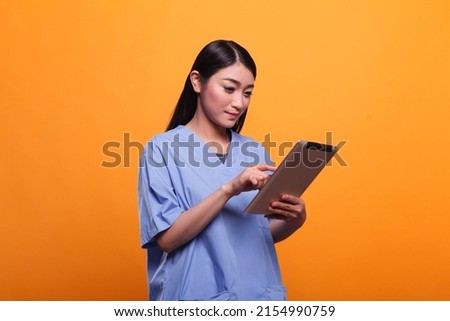 Pretty asian hospital nurse wearing blue medical uniform while using modern tablet on orange background. Clinic healthcare staff member using digital tablet. Studio shoot. Royalty-Free Stock Photo #2154990759