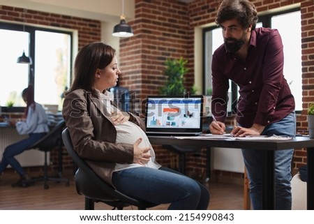 Pregnant financial company advisor being informed by employee about agency partnership revised policy. Expectant mother at work listening to colleague explaining bad results of annual revenue. Royalty-Free Stock Photo #2154990843