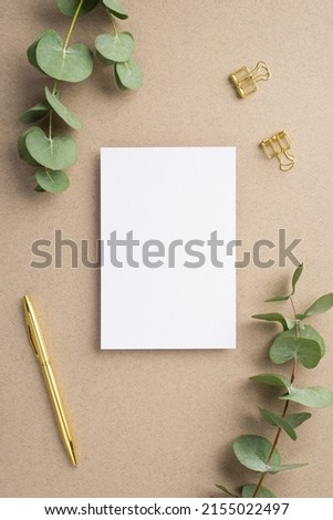 Top view vertical photo of paper card gold pen binder clips and eucalyptus on beige background with empty space
