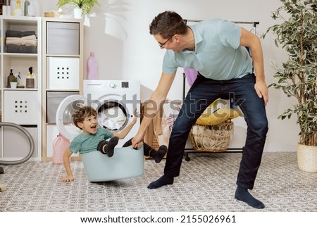 You can have fun with children, even in the bathroom, the main thing little imagination. Dad puts his son in bowl and rolls on floor. The boy laughs and waves arms. Behind it is washing machine. Royalty-Free Stock Photo #2155026961